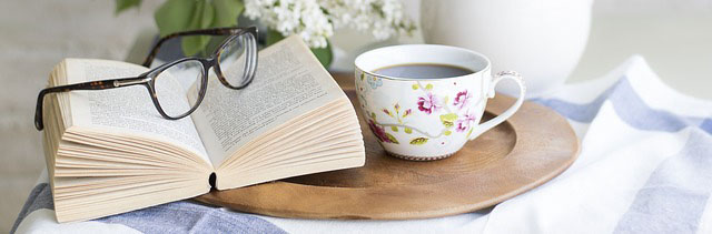 Symbolbild Lesen: aufgeschlagenes Buch neben Kaffeetasse und BVlumenvase auf einem Holztablett
