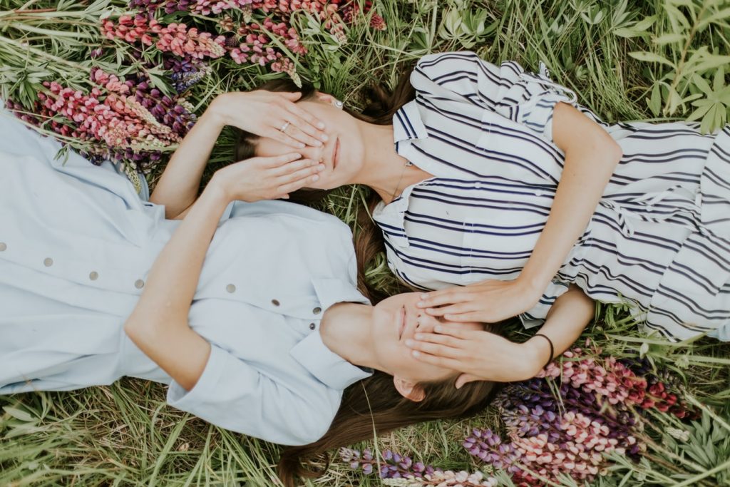 Zwillings-Frauen liegen am Rücken in einer Blumenwiese und halten sich gegenseitig die Augen zu, Credit: Daiga Ellaby, Unsplash