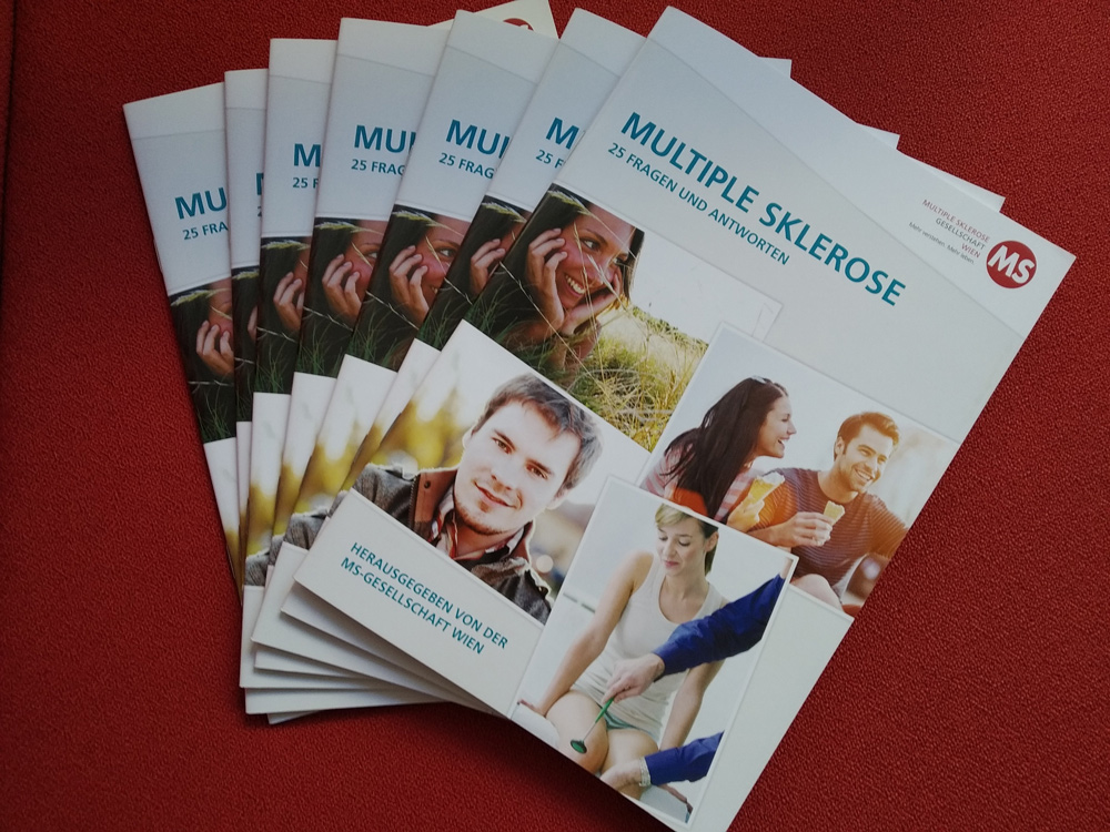 7 Ausgaben der Broschüre" Multiple Sklerose: 25 Fragen und Antworten" liegen auf einem roten Stoff. Herausgegeben von der Multiple Sklerose Gesellschaft Wien im November 2020